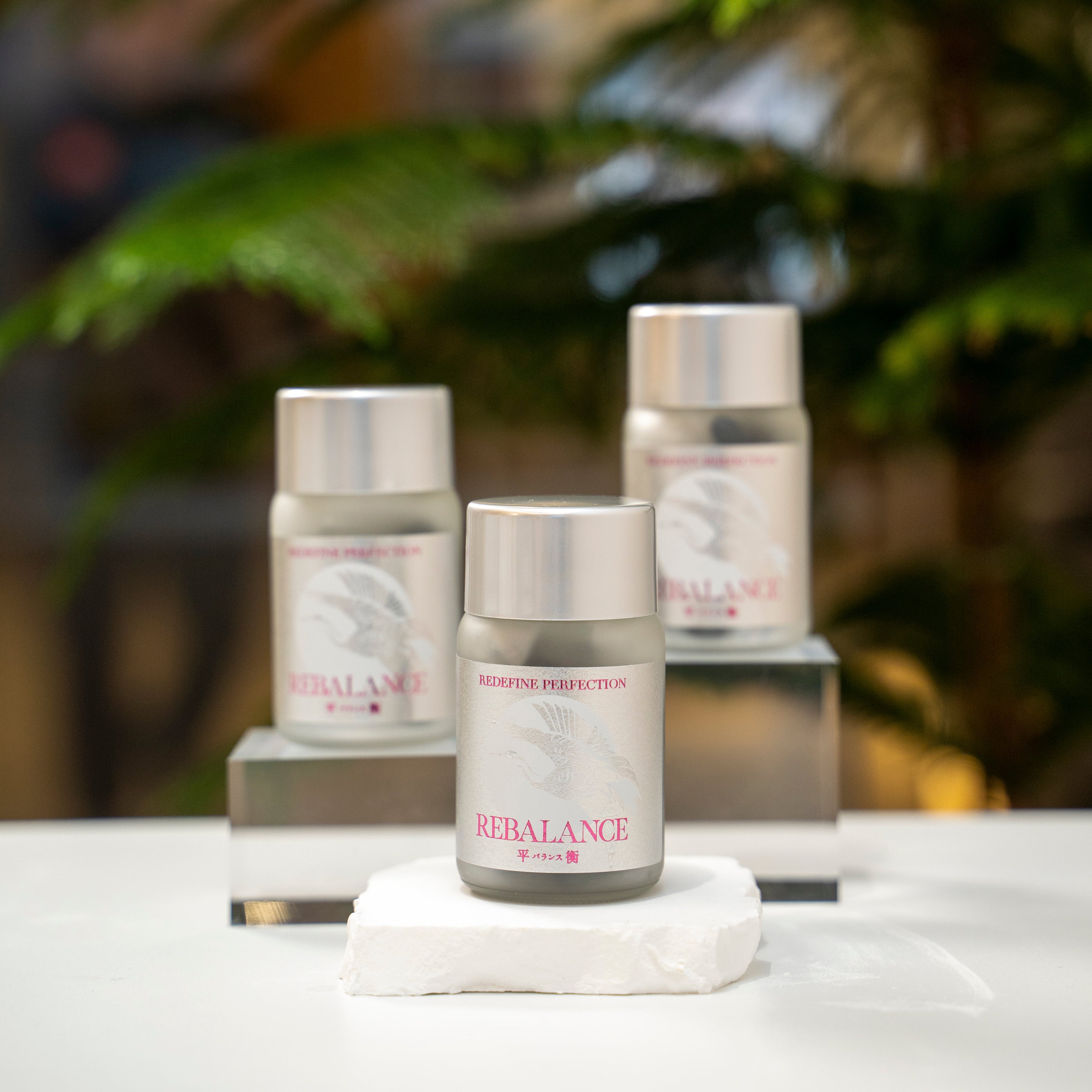Three skincare products labeled 'Rebalance' on a white surface with a blurred green plant background.
